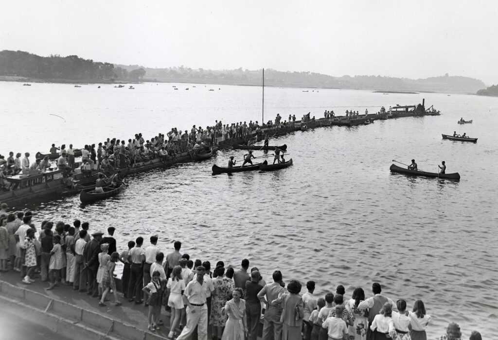 A group of onlookers surround competitors trying to tip each other off of canoes in the Susquehanna River.