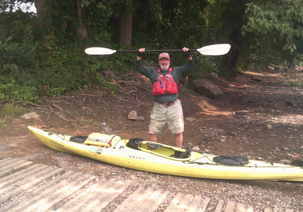 a paddler stands behind his kayak with paddle in the air