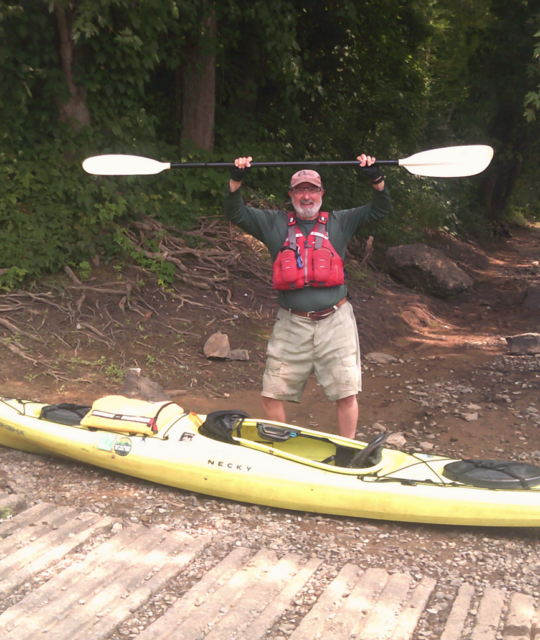 a paddler stands behind his kayak with paddle in the air
